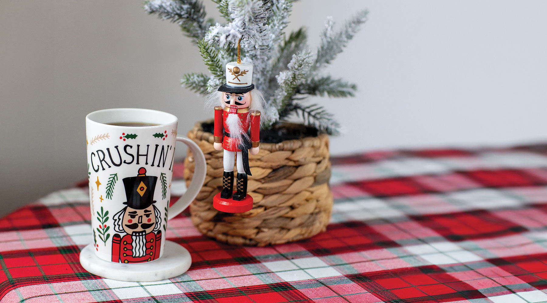 Mug with Christmas design, nutcracker figurine, and small tree on a plaid tablecloth.