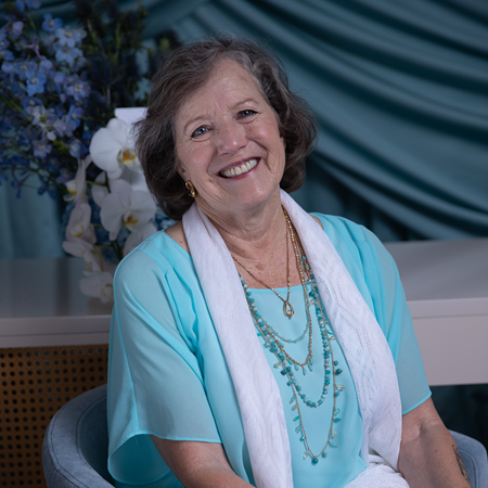 Artist Sharon Thayer wearing a light blue top and white scarf with layered necklaces, sitting in a room with decorative plants.